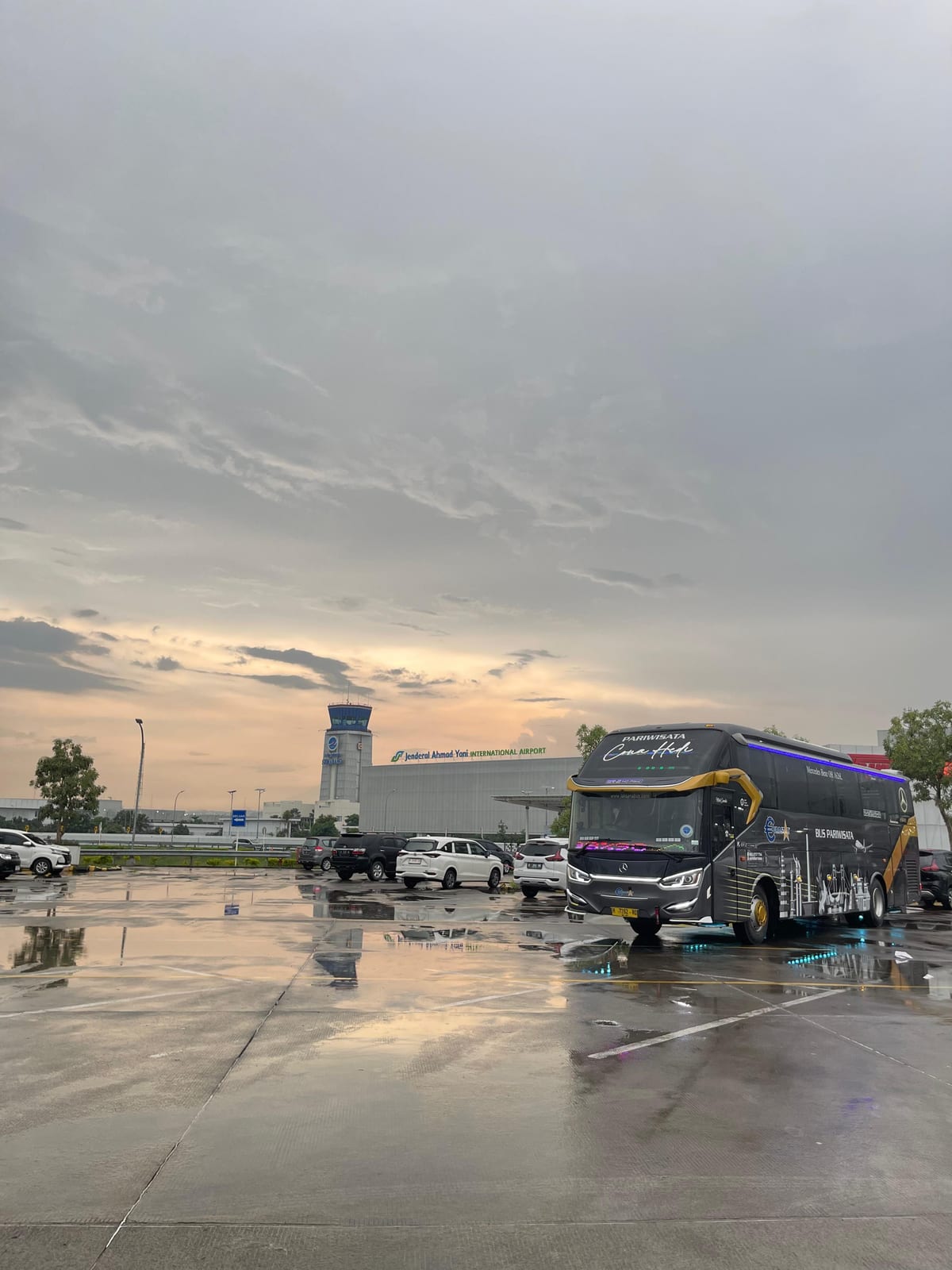 Bus Cona Hede menunggu di area parkir Bandara Internasional Yogyakarta saat senja setelah hujan
