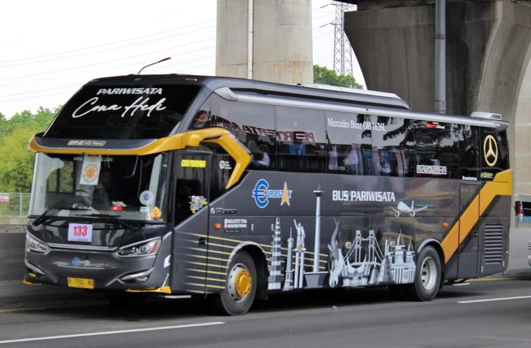 Bus Cona Hede berlivery abu-abu gelap dan emas melaju di jalan raya, tampak samping depan