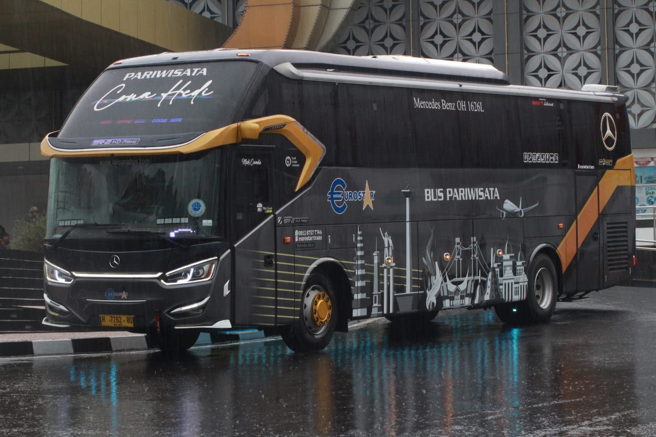 Bus pariwisata Mercedes-Benz OH 1626L Cona Hede berwarna abu-abu gelap dengan aksen emas, tampak samping depan di area gedung saat hujan
