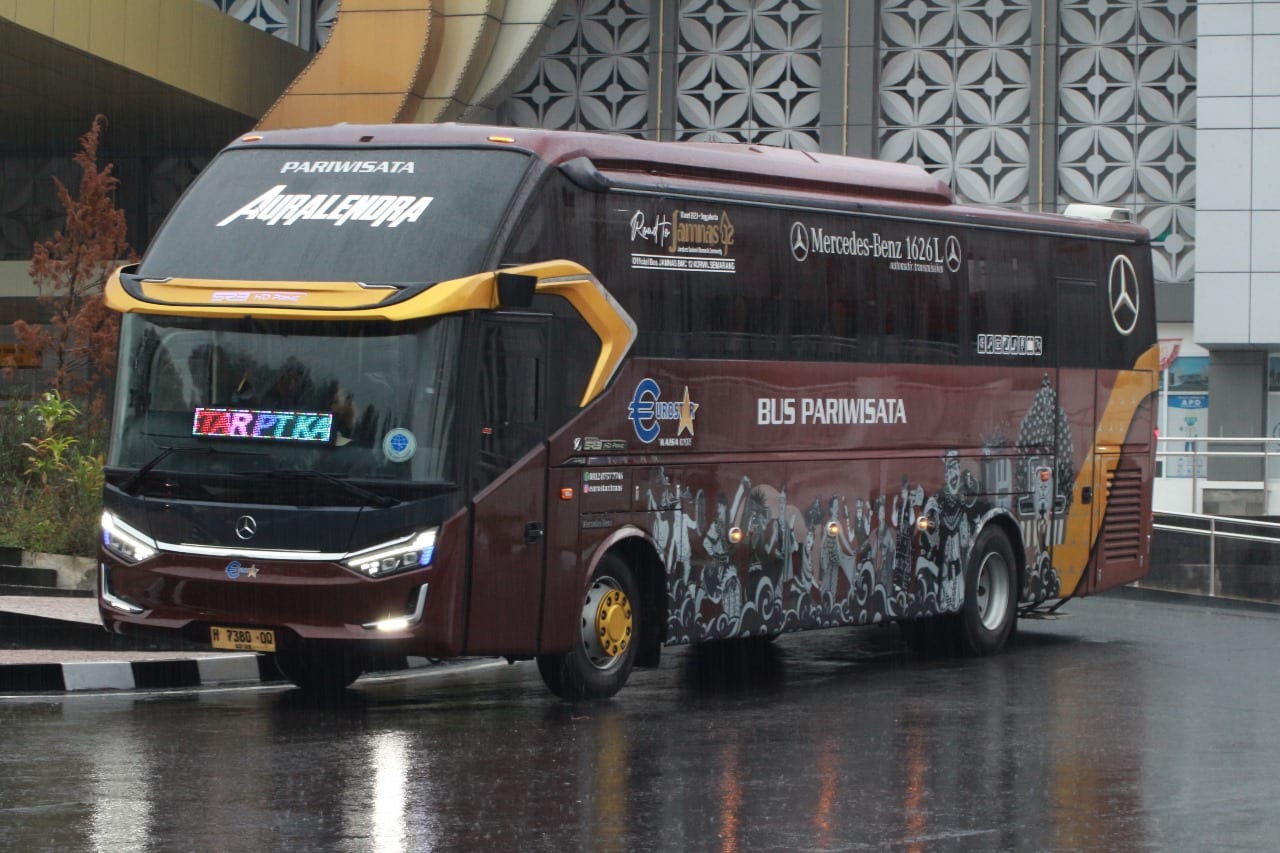 Bus pariwisata Auralendra berwarna marun dan emas, tampak samping depan di area gedung saat hujan