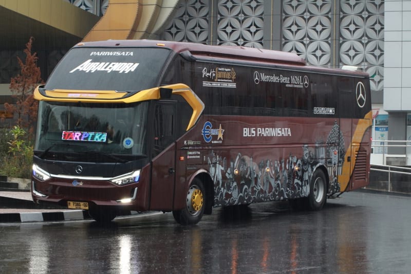 Bus pariwisata Auralendra berwarna marun dan emas, tampak samping depan di area gedung saat hujan