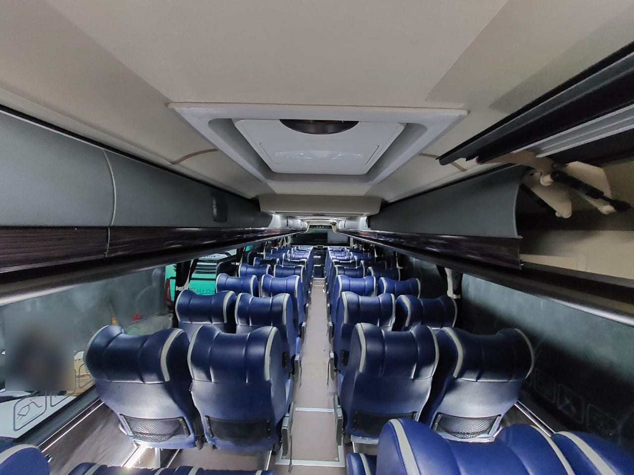Kabin bus dengan deretan kursi biru formasi dua-dua dan bagasi kabin di atas kursi