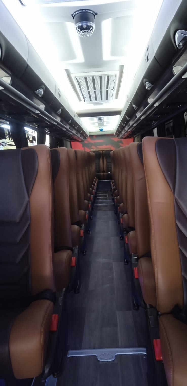Lorong kabin bus dengan deretan kursi kulit berwarna cokelat di kedua sisi dan lampu kabin menyala